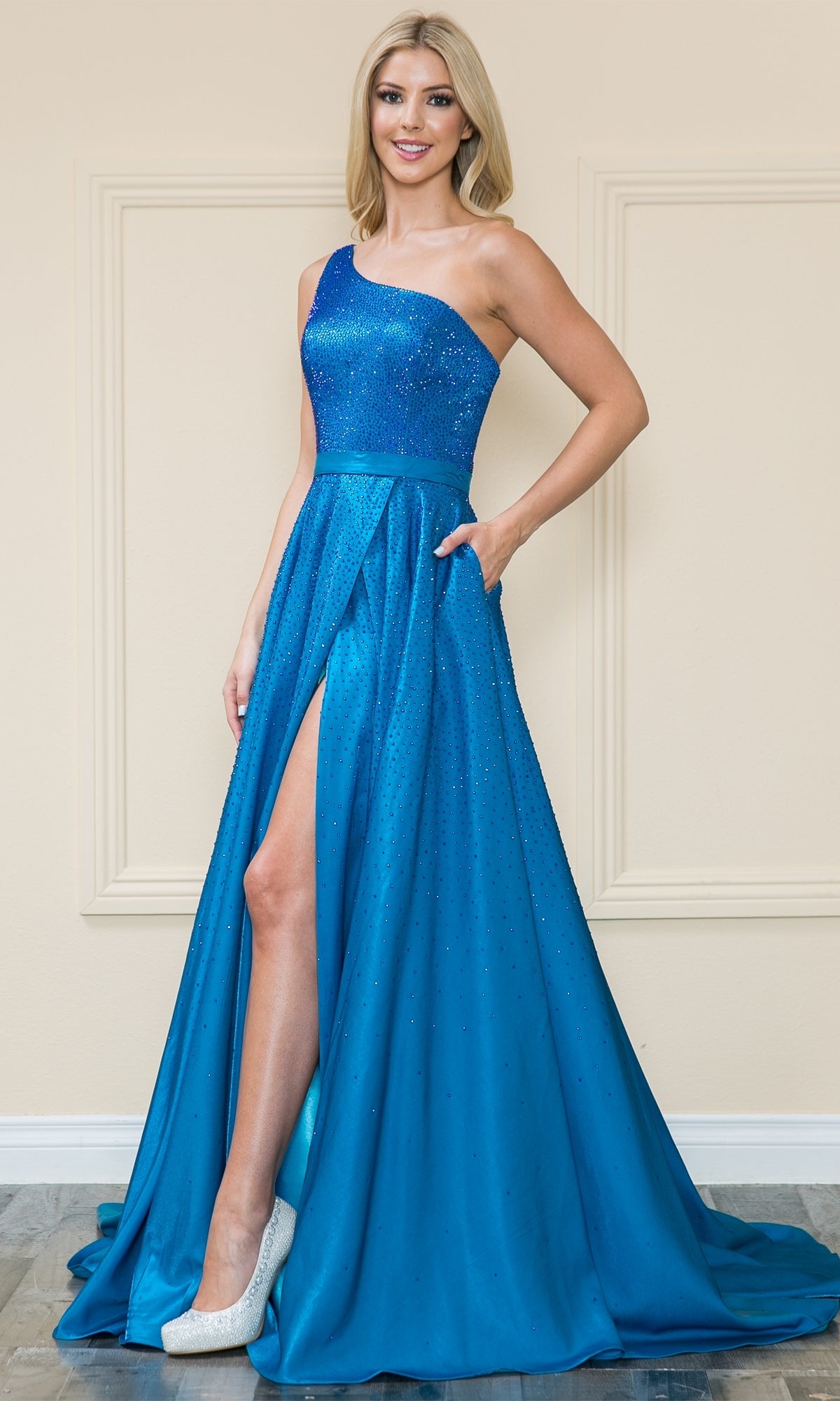 A woman stands indoors wearing the Poly USA 8914 One-Shoulder Long A-Line Prom Gown in vibrant blue, featuring sparkling beaded details, a high slit, and paired with white high-heeled shoes.